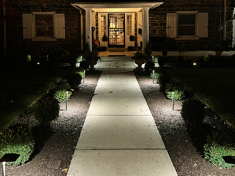 Front walkway in Overbrook Farms illuminated with integrated landscape lighting along the pathway and surrounding planting beds at night.