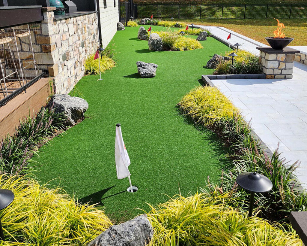 Angled view of putting green with surrounding perennial plantings, pool, and detached gym at a modern farmhouse landscape in Christiana, Pennsylvania